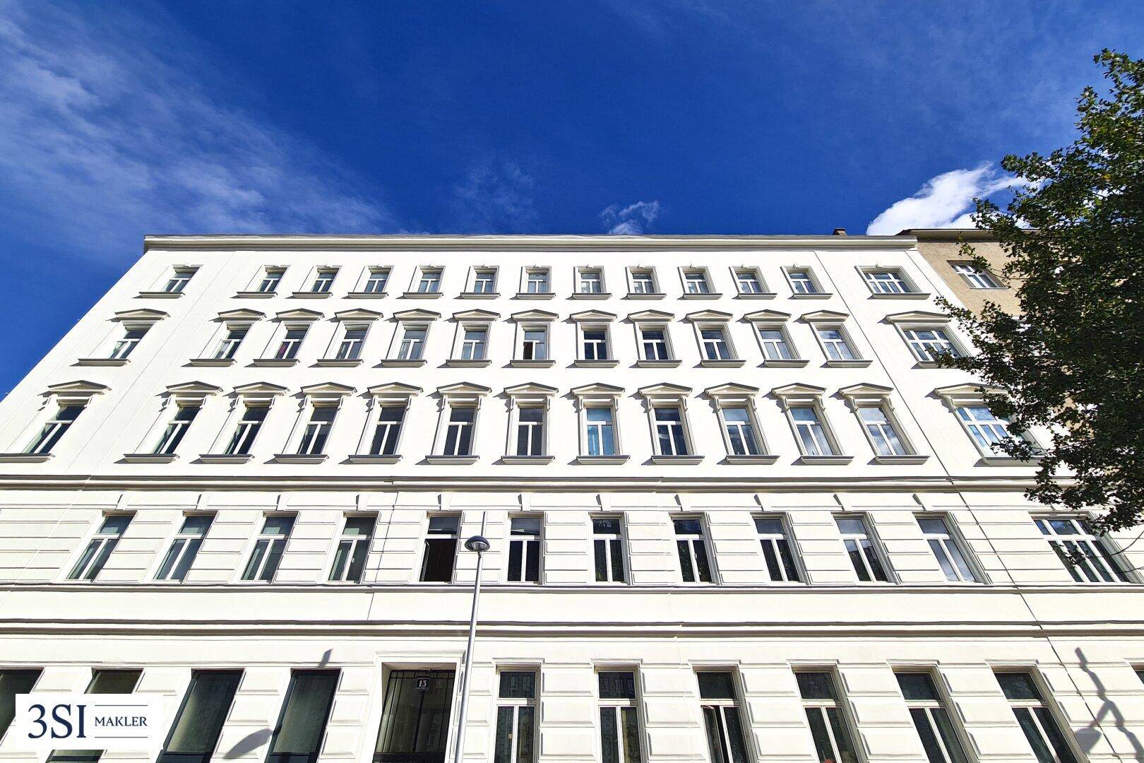 Die weiße Fassade des Mehrfamilienhauses mit vielen Fenstern unter blauem Himmel.
