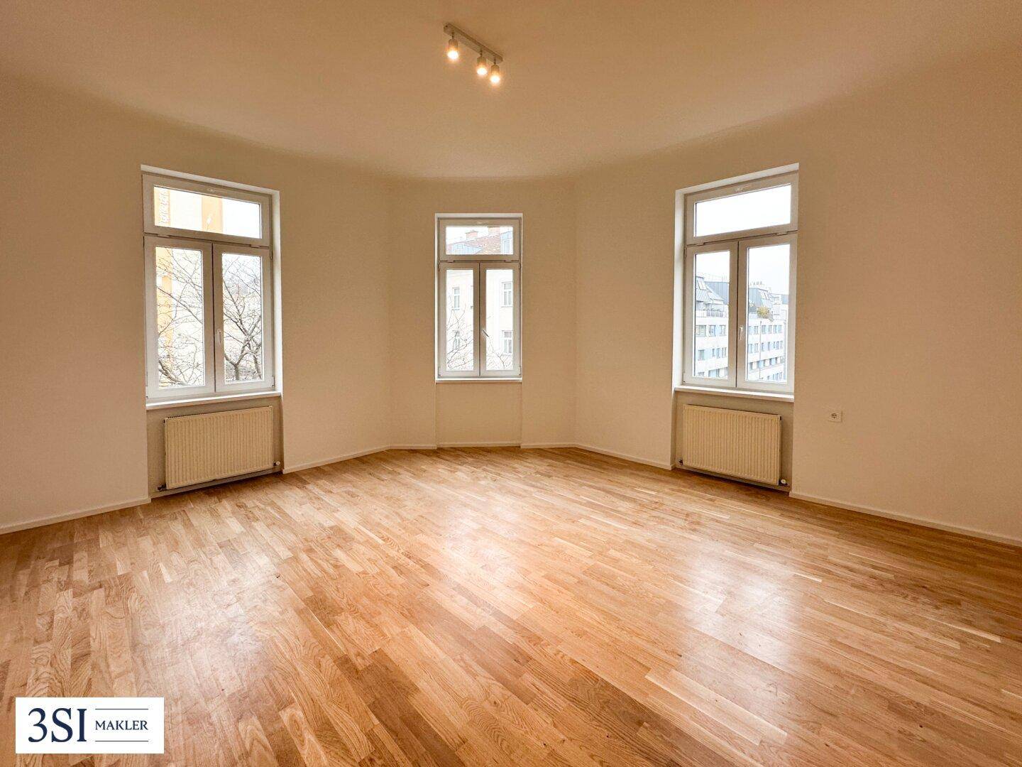 Geräumiges, leeres Wohnzimmer mit hellem Parkettboden und großen Fenstern, die den Raum lichtdurchfluten.