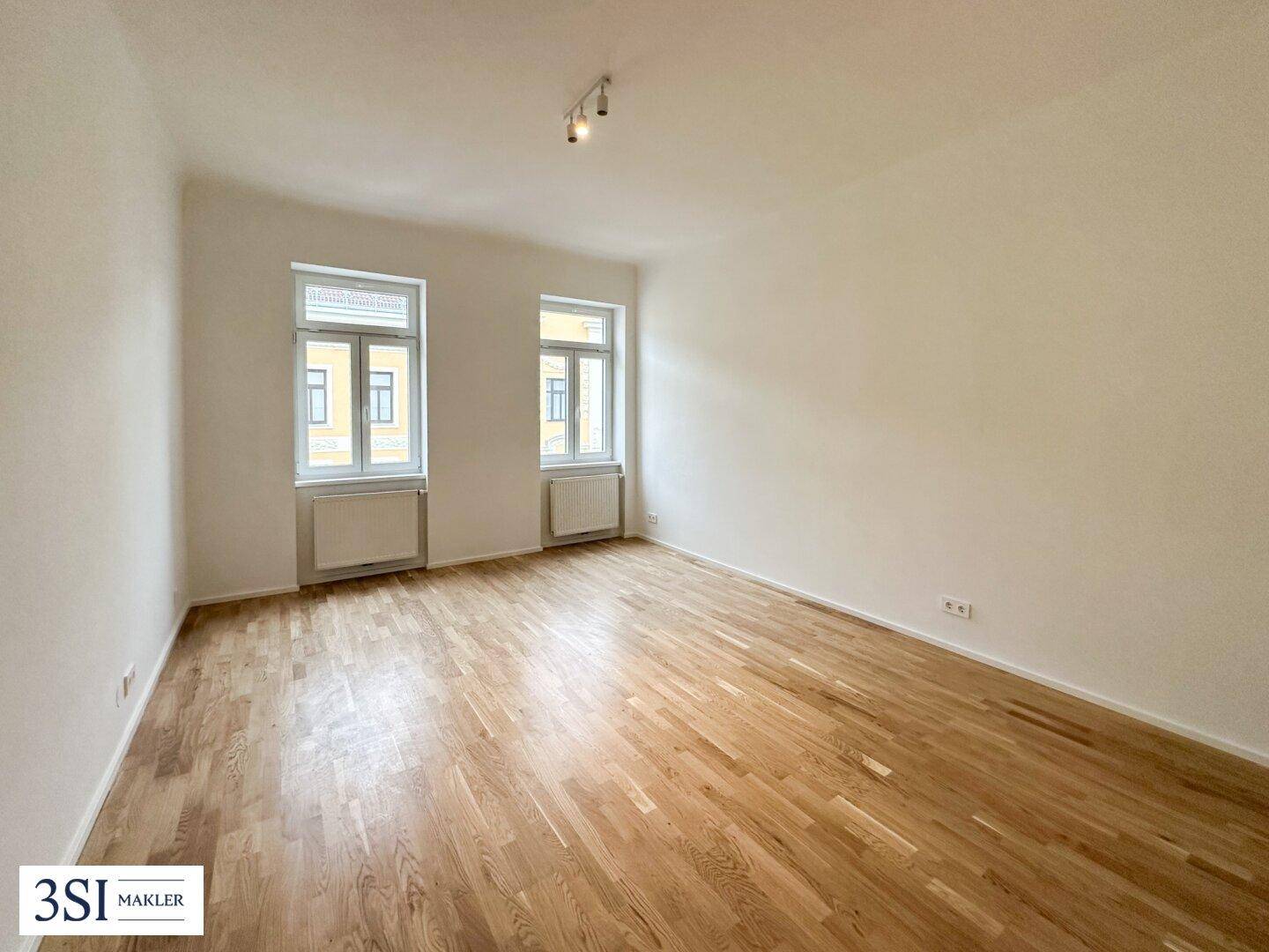 Geräumiges, leeres Zimmer mit hellem Parkettboden und zwei großen Fenstern, bereit zur individuellen Gestaltung.