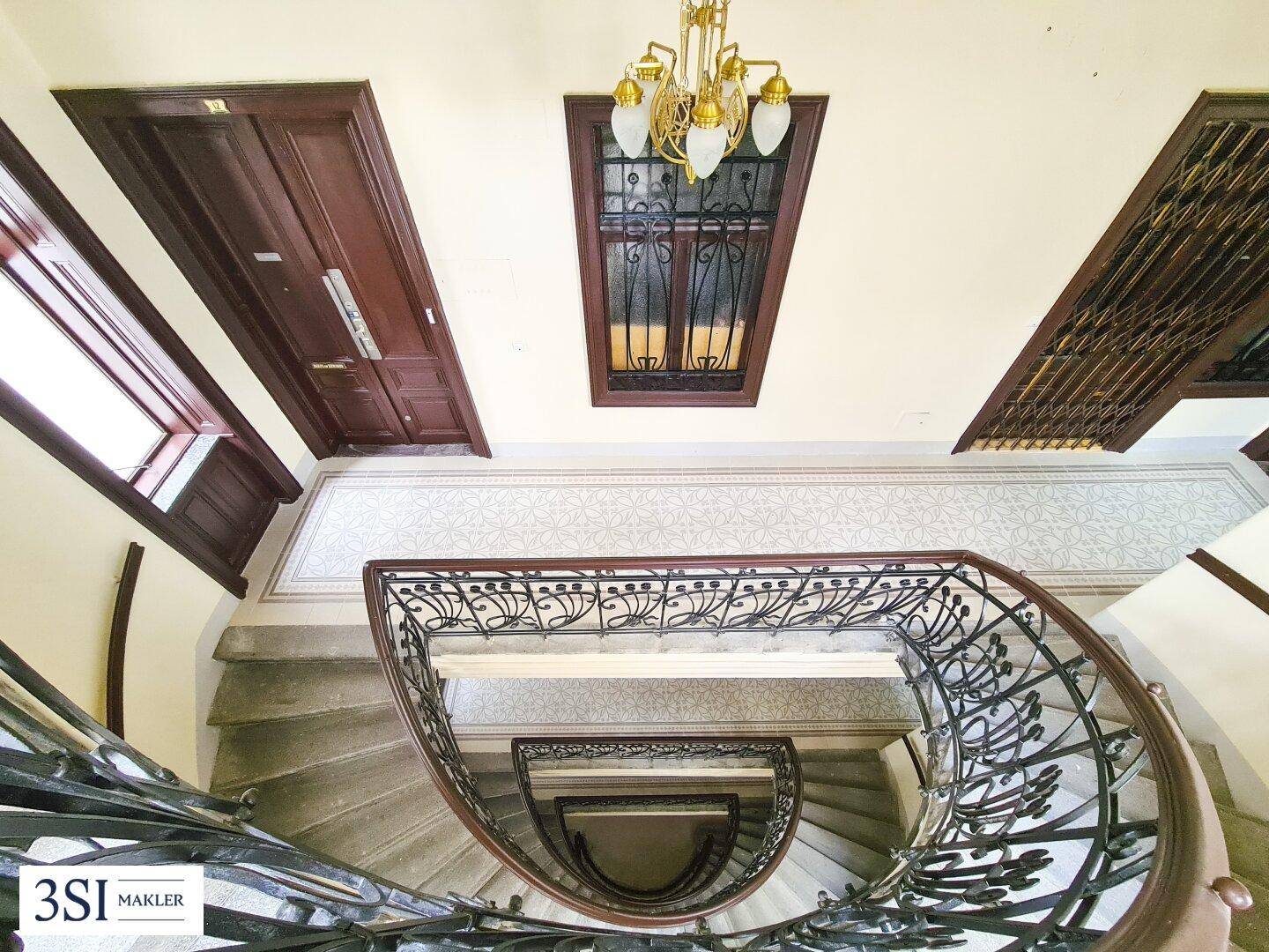Blick von oben auf ein elegantes Treppenhaus mit schmiedeeisernem Geländer und gemusterten Fliesen.