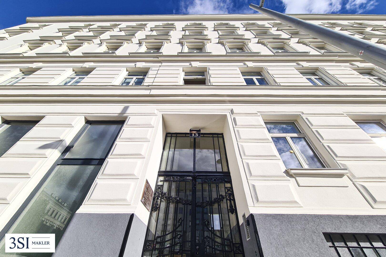 Eingangsbereich des Altbaus mit schmiedeeisernem Tor und klassischer Fassadengestaltung unter blauem Himmel.