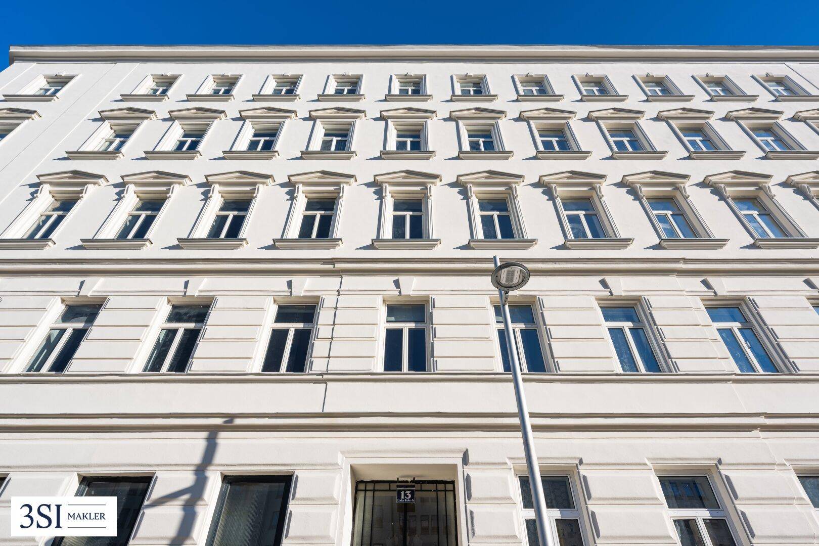 Die weiße Fassade eines Altbaugebäudes mit vielen Fenstern und dem Eingangsbereich mit Hausnummer 13.