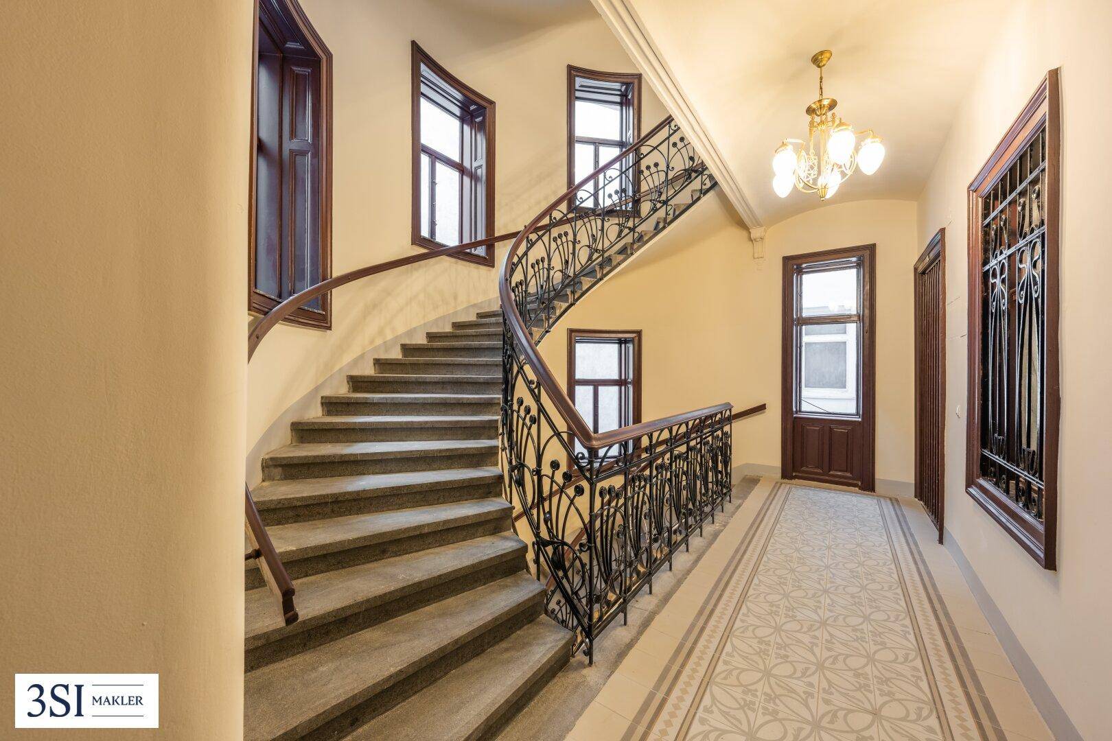 Elegantes Treppenhaus in einem Altbau mit geschwungenem Geländer, gemustertem Boden und hohen Decken.