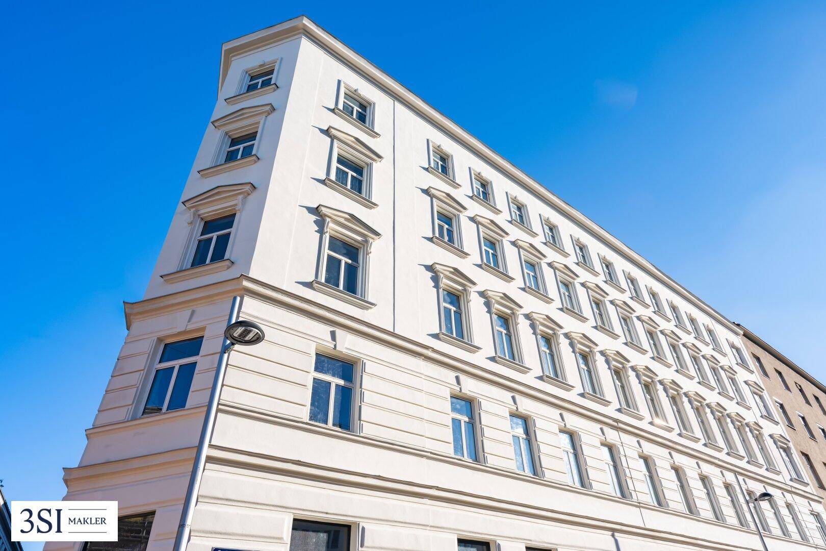 Die helle Fassade eines Eckgebäudes mit vielen Fenstern unter strahlend blauem Himmel.