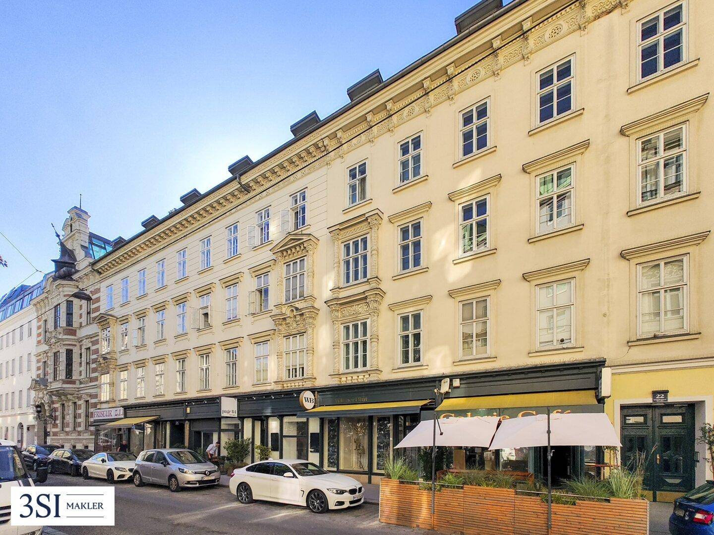 Lange Reihe von Altbauten entlang einer Straße mit parkenden Autos und Sonnenschirmen vor Geschäften.