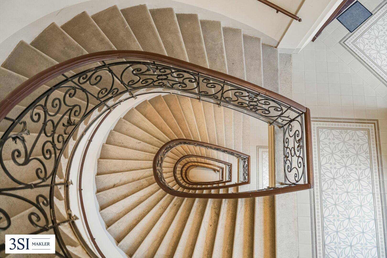 Elegantes Treppenhaus mit geschwungenem schmiedeeisernem Geländer und Teppichstufen in einem Altbau.