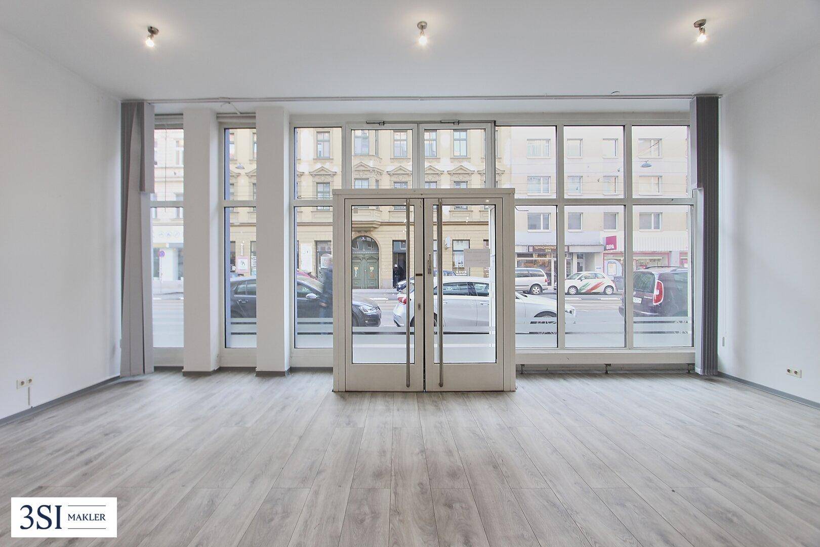 Heller, leerer Gewerberaum im Erdgeschoss mit großen Schaufenstern zur Straße und modernem Laminatboden.