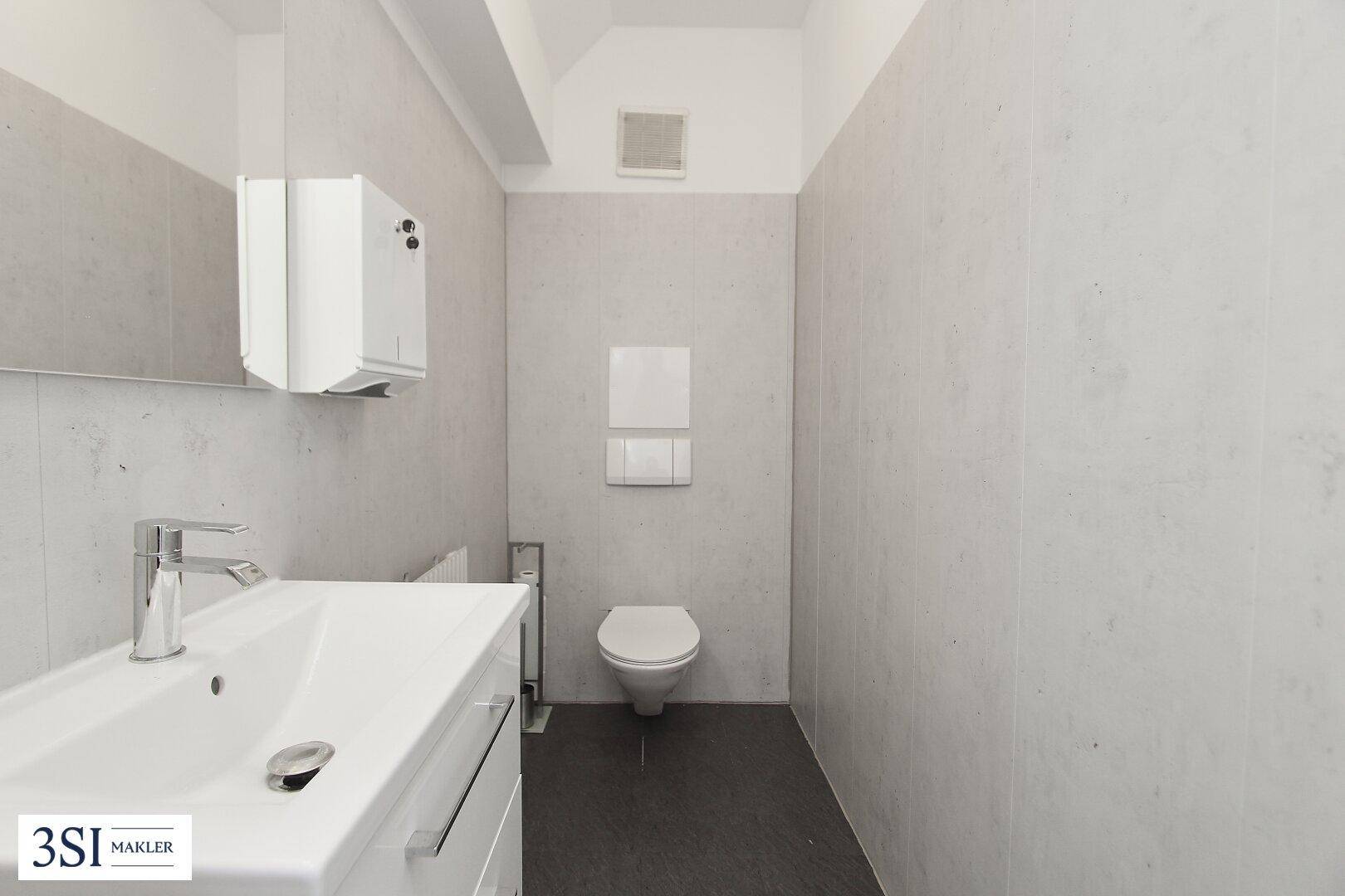 Modernes WC mit Waschbecken, Wand-WC und hellgrauen Fliesen an Wänden sowie dunklem Boden.