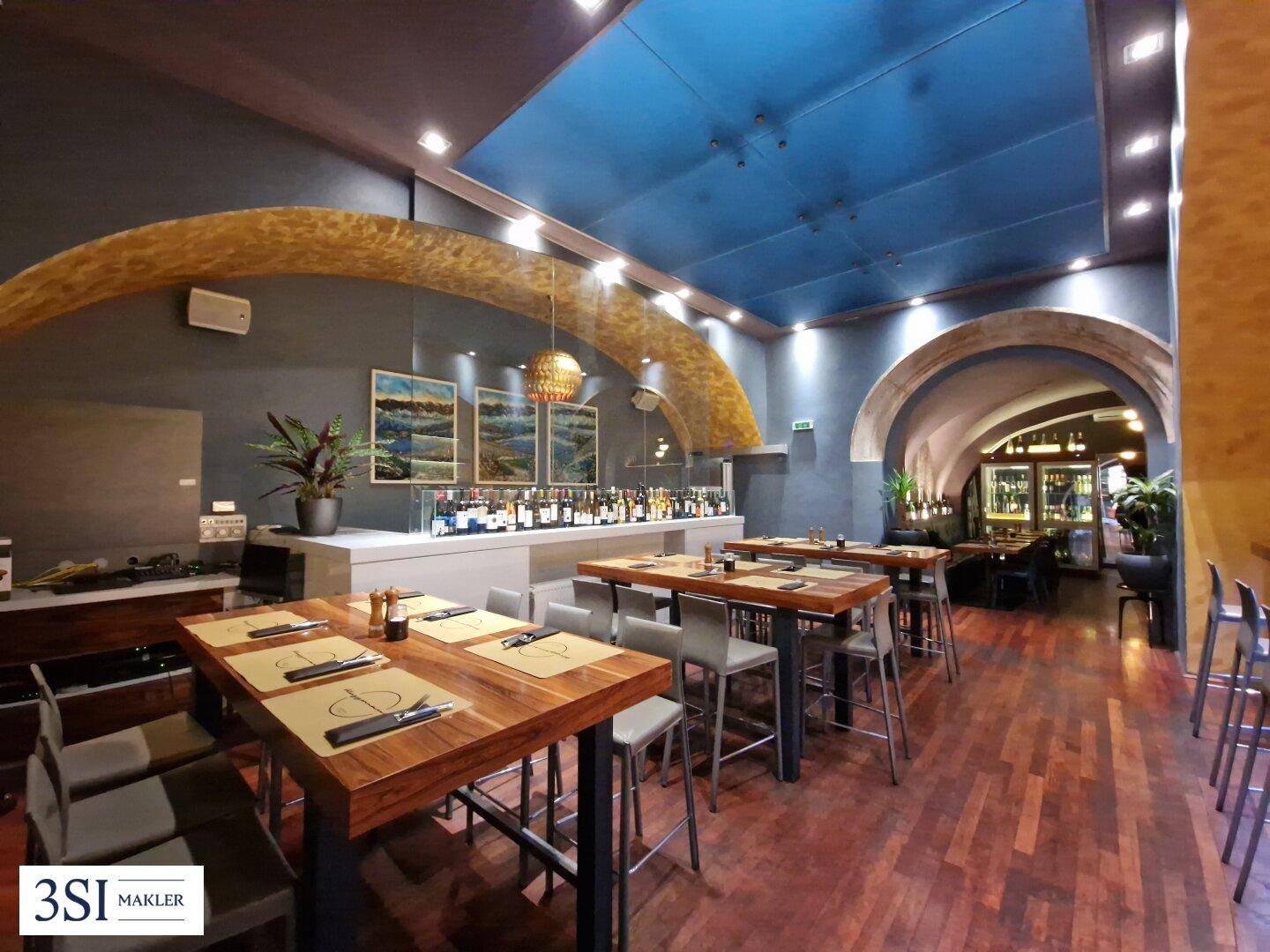 Moderner Restaurantbereich mit Holztischen, Bar und blauer Deckenbeleuchtung.