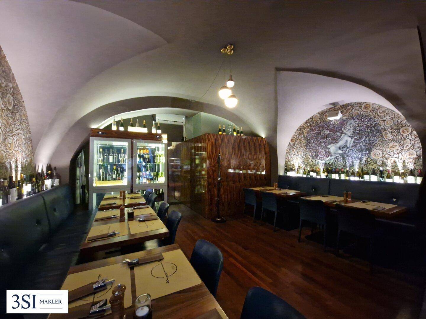 Langer Restaurantraum mit Holztischen, Sitzbänken und integrierten Weinkühlern an der Wand.