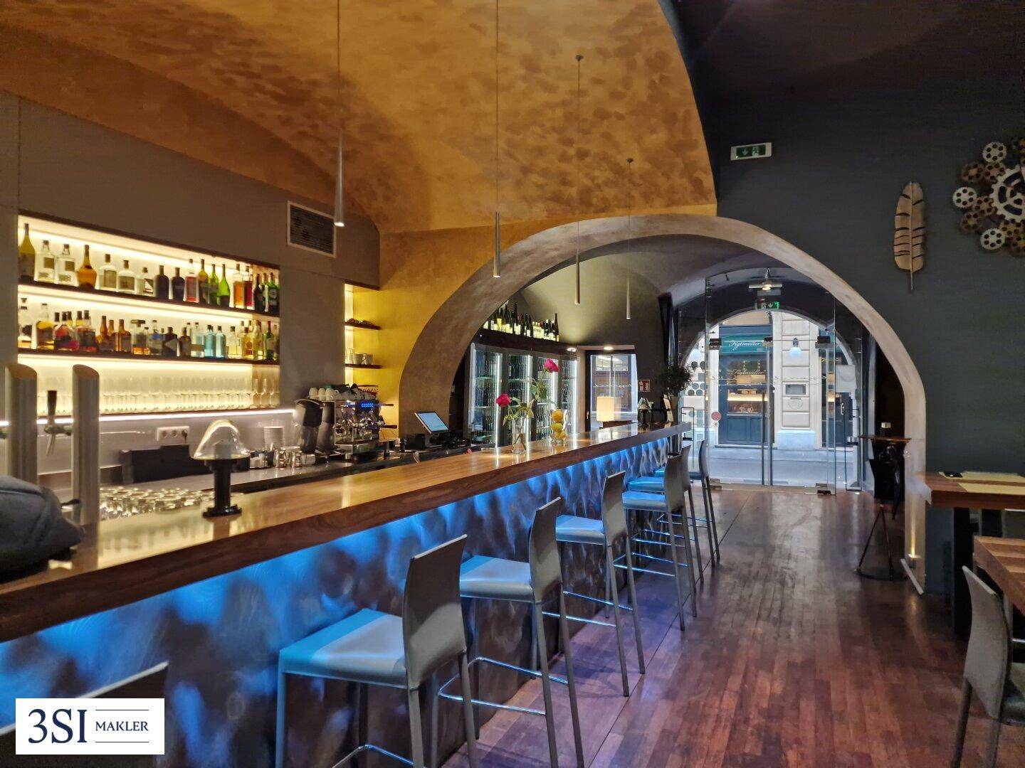 Moderne Bar mit beleuchteter Theke, hohen Hockern und Blick zum historischen Eingangsbereich.