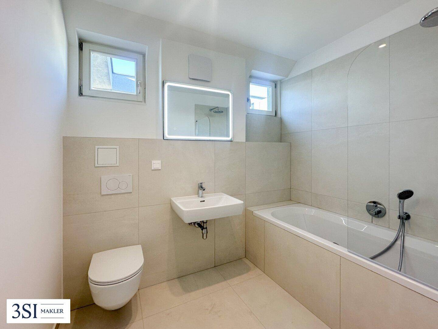 Modernes Badezimmer mit Badewanne, Wand-WC, Waschbecken und zwei Fenstern für gute Belüftung und Licht.