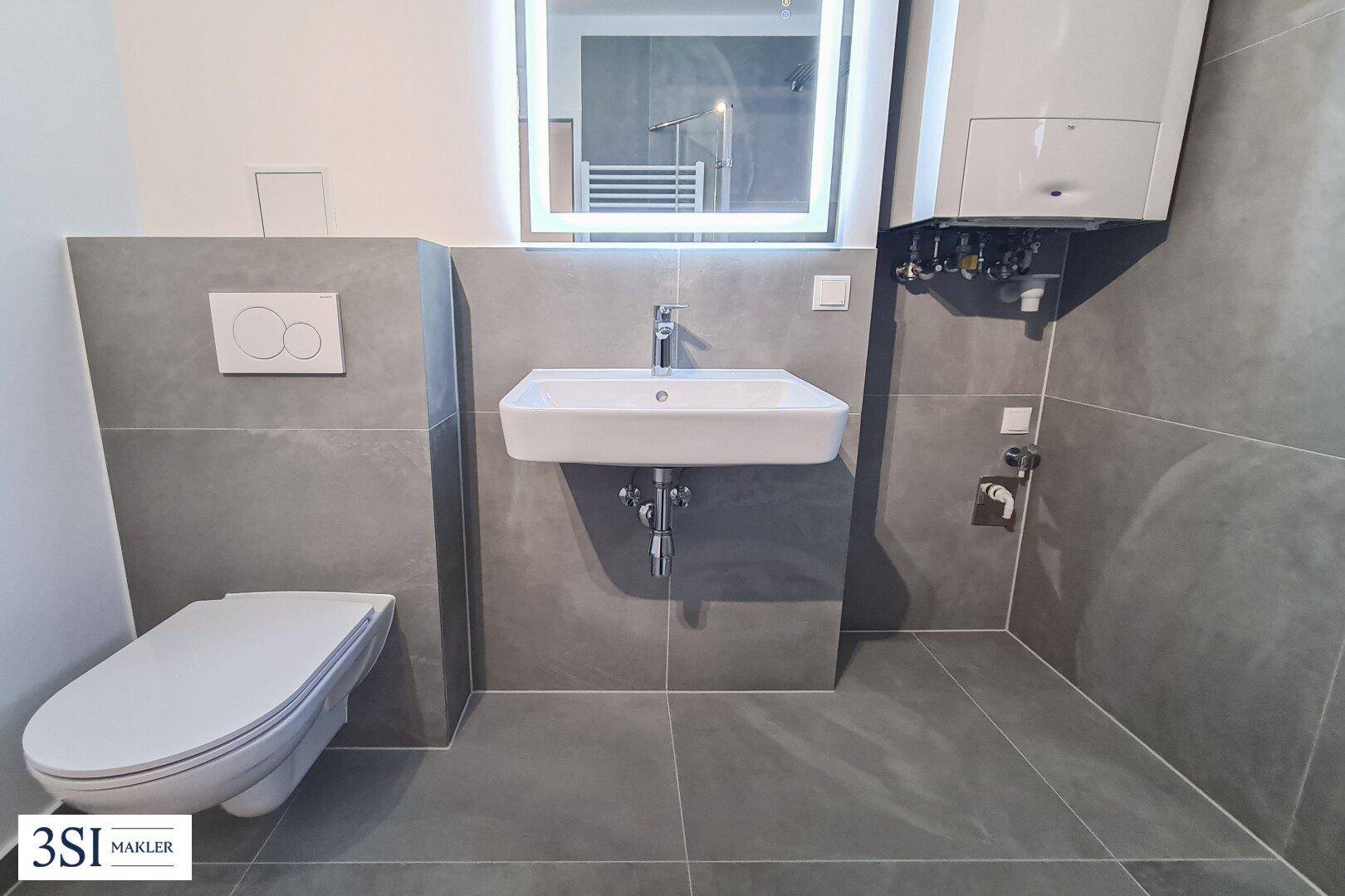Elegantes Badezimmer mit grauen Fliesen, modernem Waschtisch und beleuchtetem Spiegelschrank.