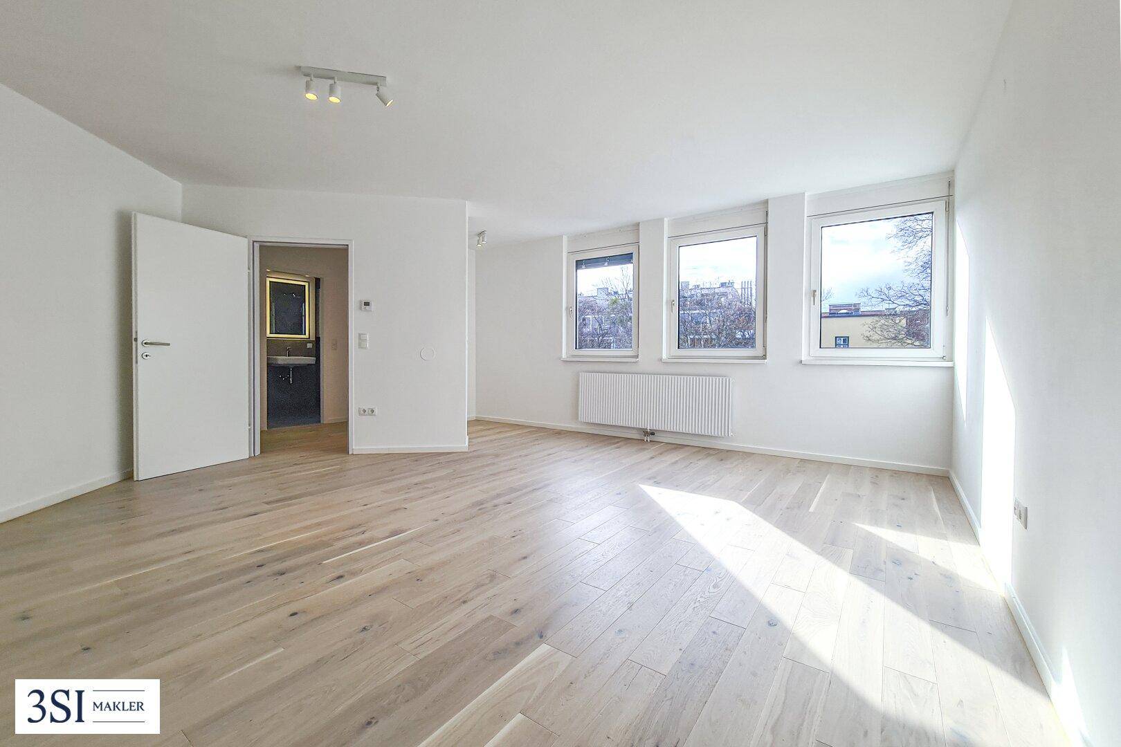 Lichtdurchfluteter Wohnraum mit Holzboden, großen Fenstern und offenem Zugang zu einem angrenzenden Badezimmer.