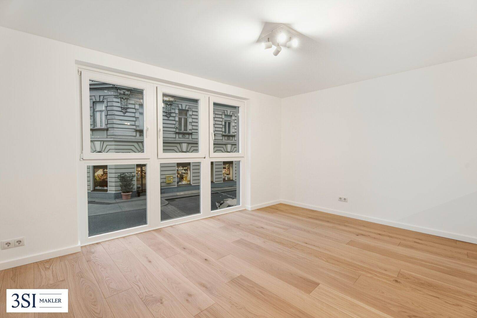 Helles, leeres Zimmer mit Parkettboden und großen Fenstern zur belebten Straße.