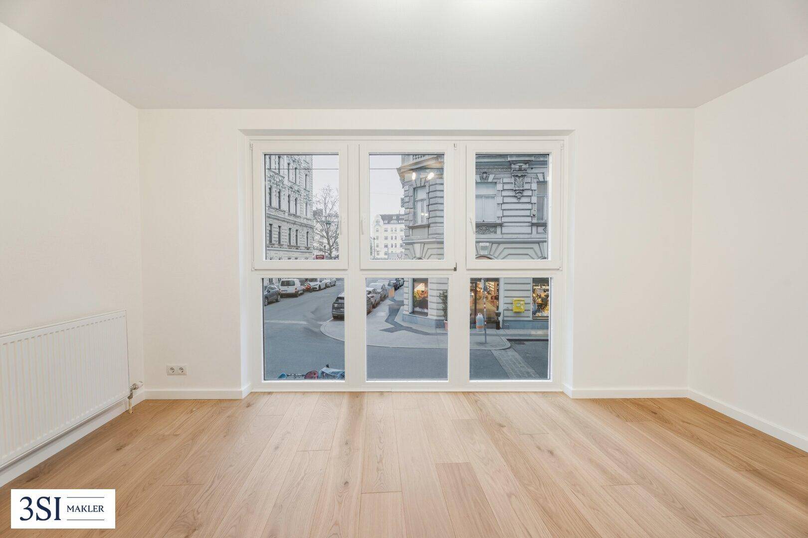 Helles, leeres Zimmer mit Parkettboden und großen Fenstern zur belebten Straße.