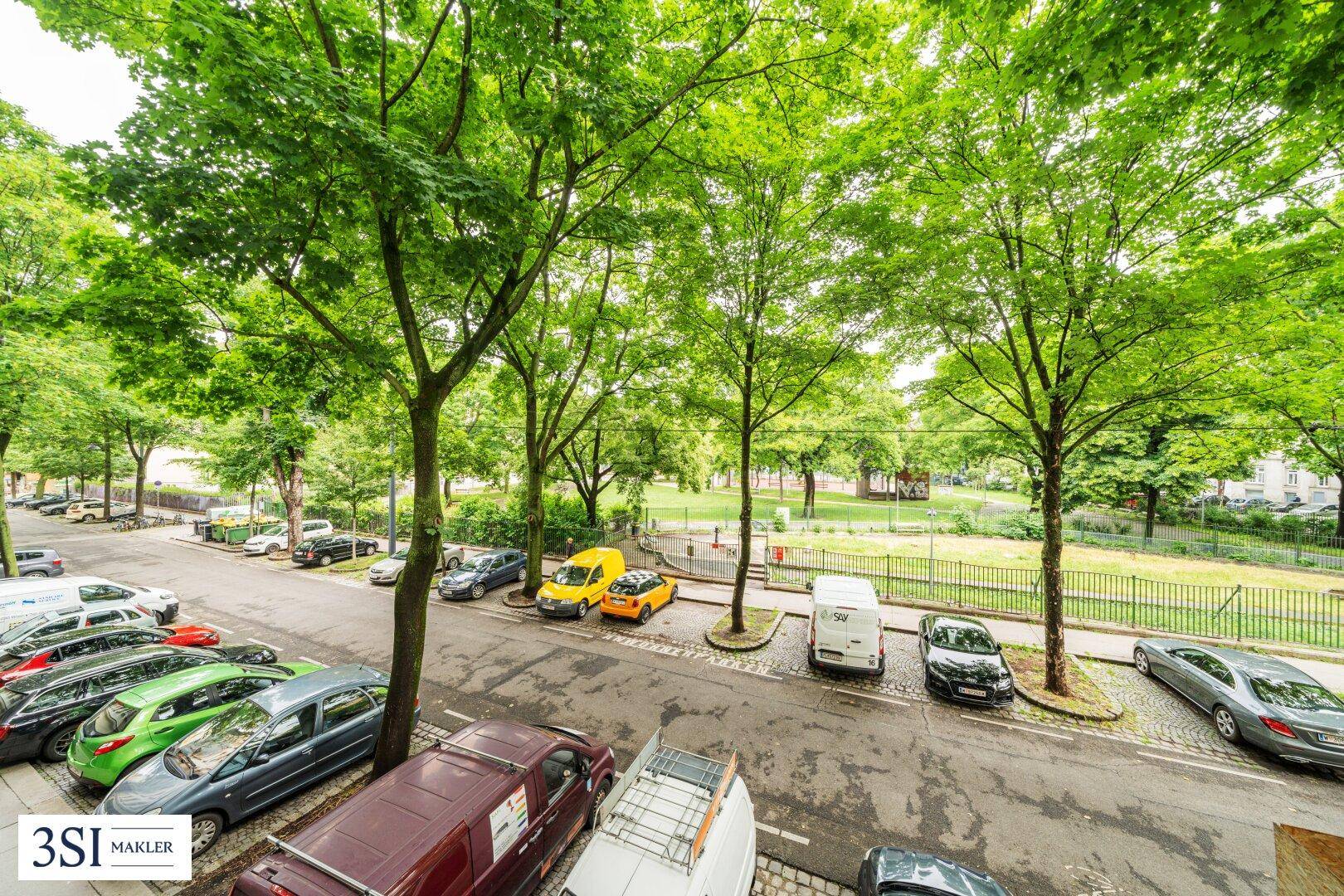Blick auf eine belebte Straße mit parkenden Autos und einem angrenzenden grünen Parkbereich.