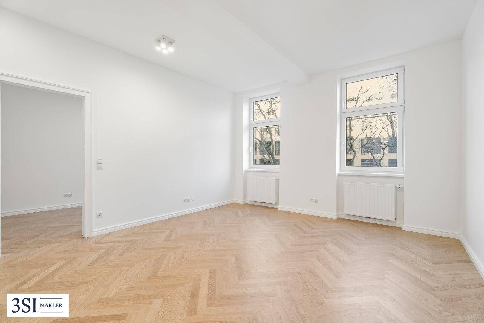 Lichtdurchflutetes Zimmer mit Fischgrätparkett, weißen Wänden und Blick ins Grüne.