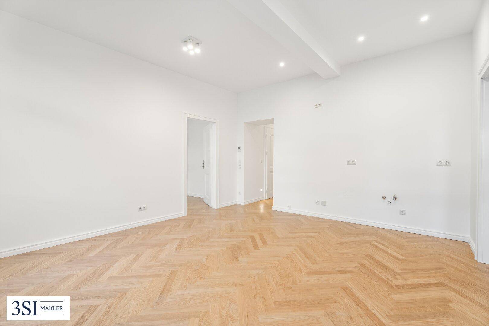 Geräumiges Zimmer mit Fischgrätparkett, weißen Wänden und Zugang zu weiteren Räumen.