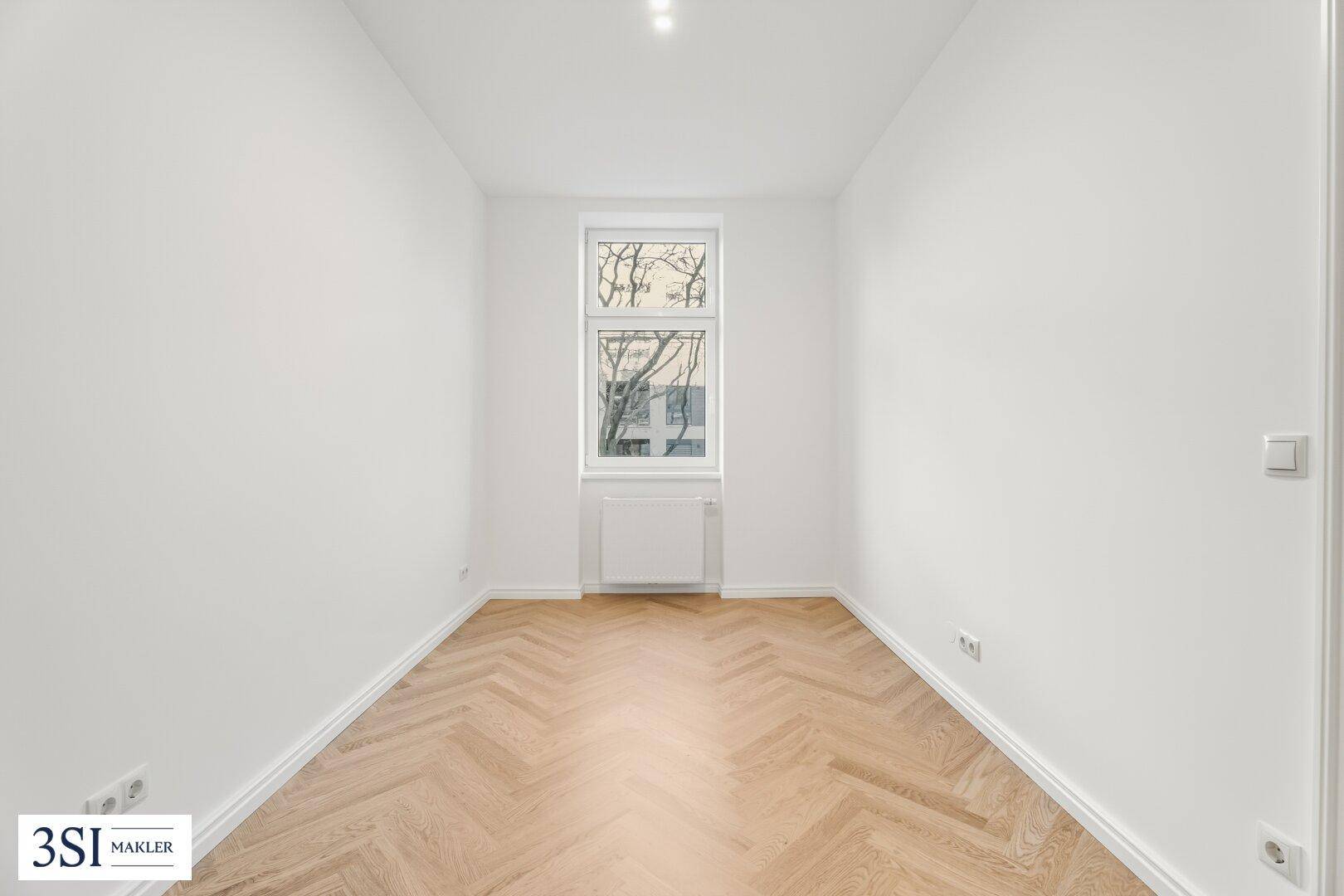 Schmaler Raum mit Fischgrätparkett, weißen Wänden und einem Fenster, ideal als Schlafzimmer.