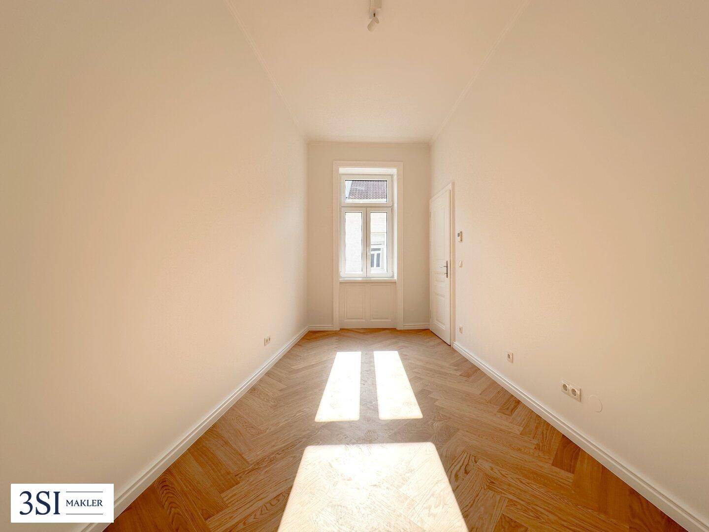 Langes, helles Zimmer mit Fischgrätparkettboden und einem großen Fenster, das viel Licht hereinlässt.