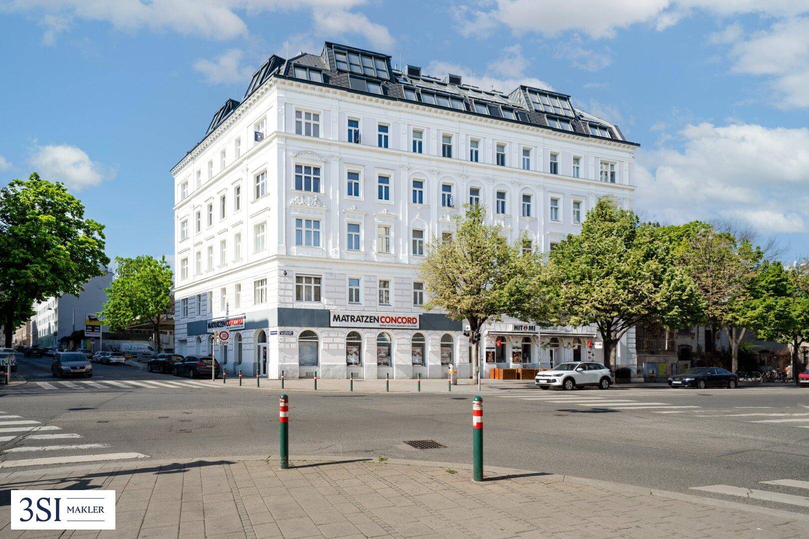 Klassisches Wohngebäude an einer Straßenecke mit Geschäften im Erdgeschoss und Bäumen.
