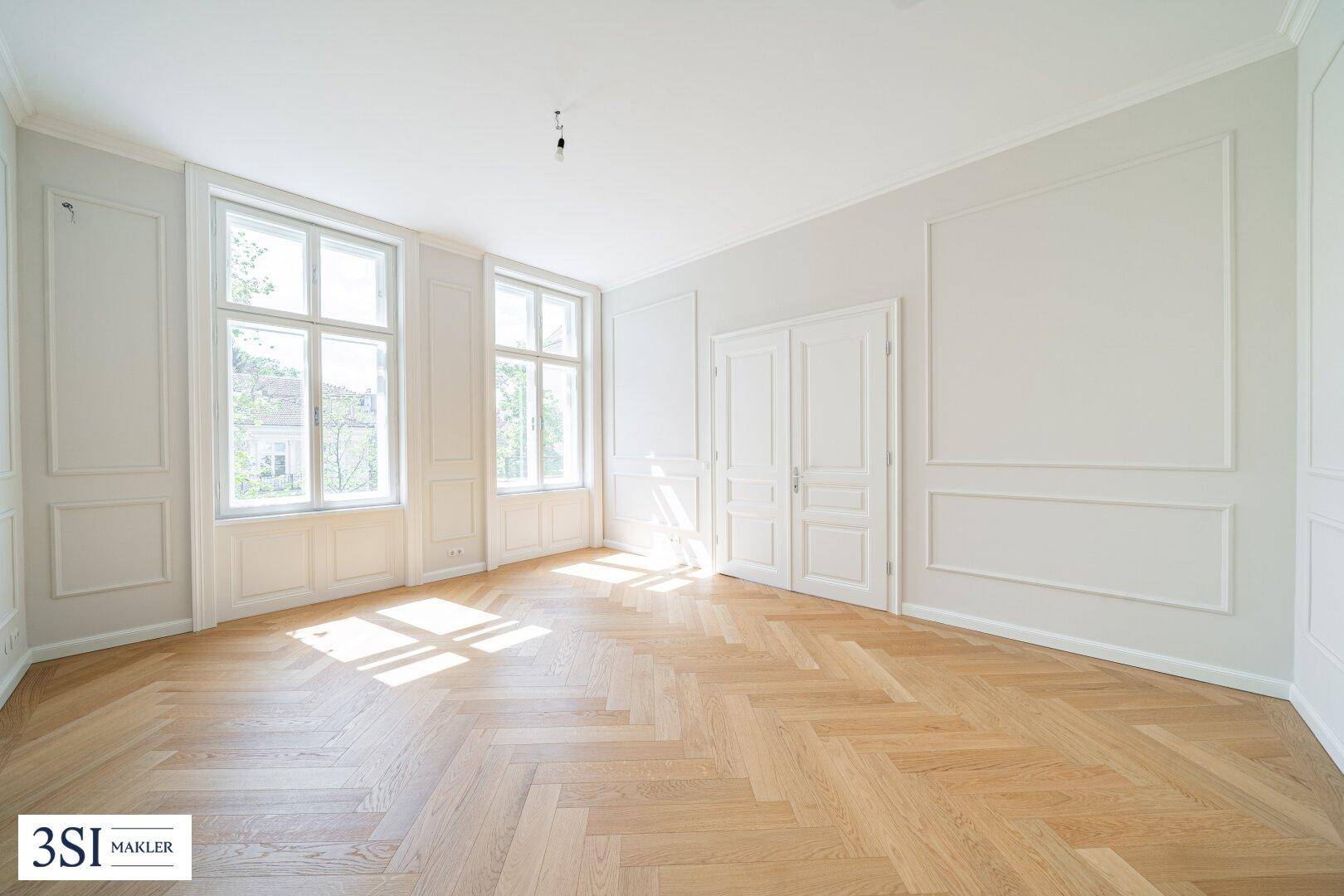 Helles Zimmer mit Fischgrätparkett, hohen Fenstern und klassischen Wandverkleidungen.