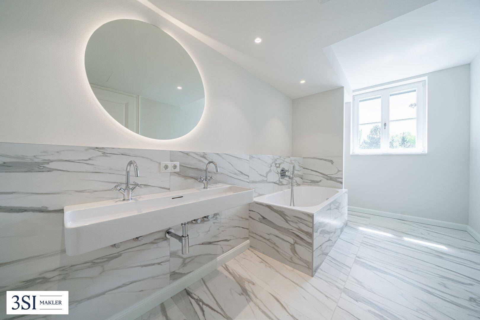 Elegantes Badezimmer mit Marmoroberflächen, Doppelwaschbecken und beleuchtetem Spiegel.