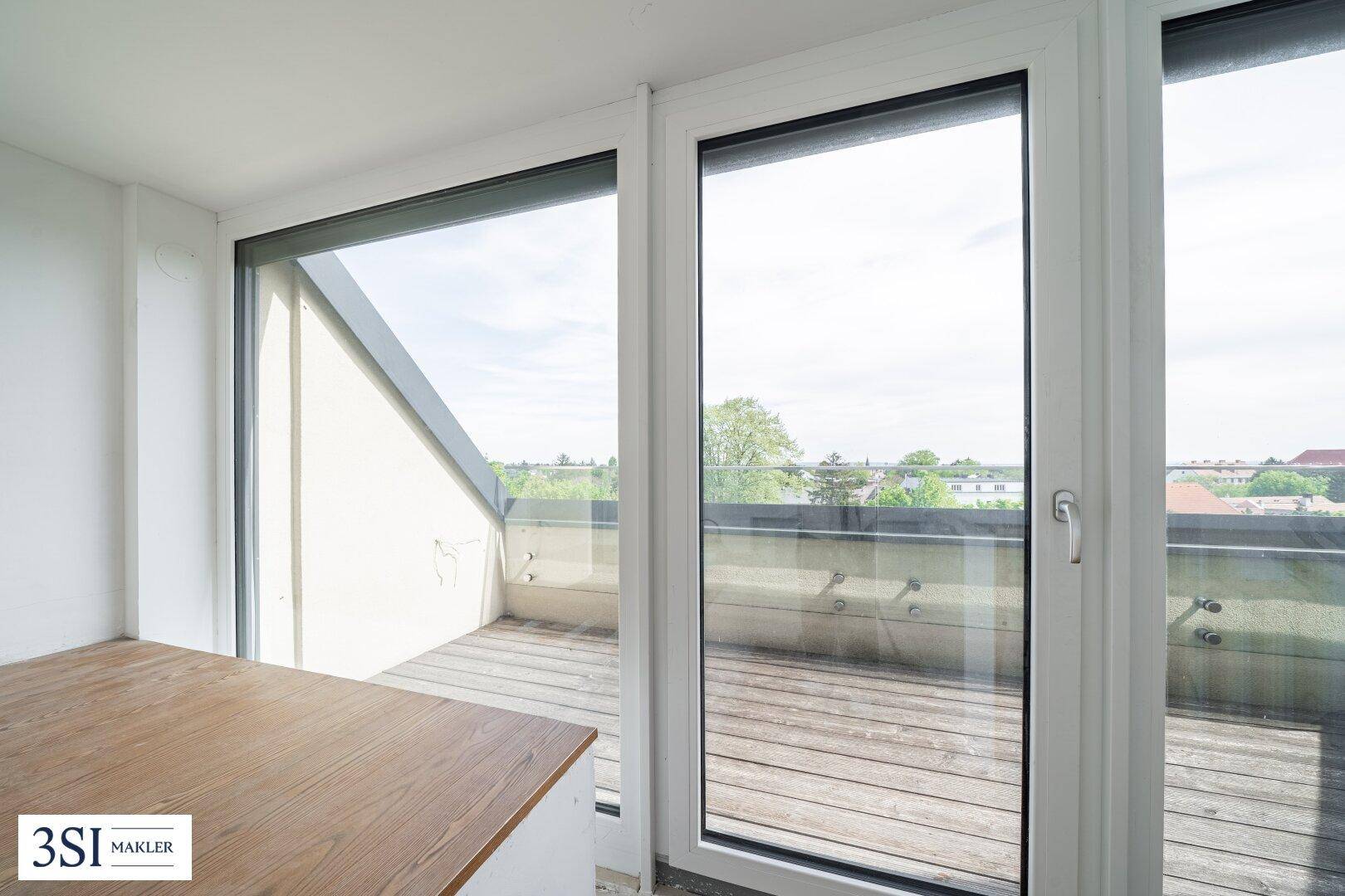 Blick von einem Innenraum durch große Fenster auf eine Holzterrasse mit Glasgeländer.