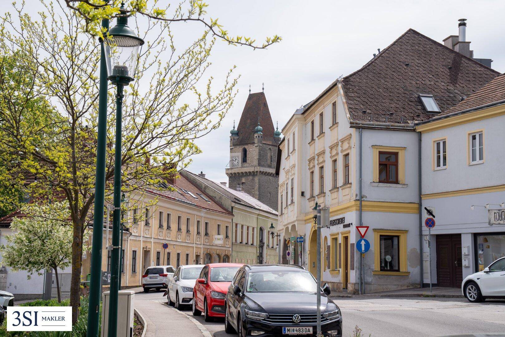 Historische Straße mit geparkten Autos und einem Kirchturm im Hintergrund.