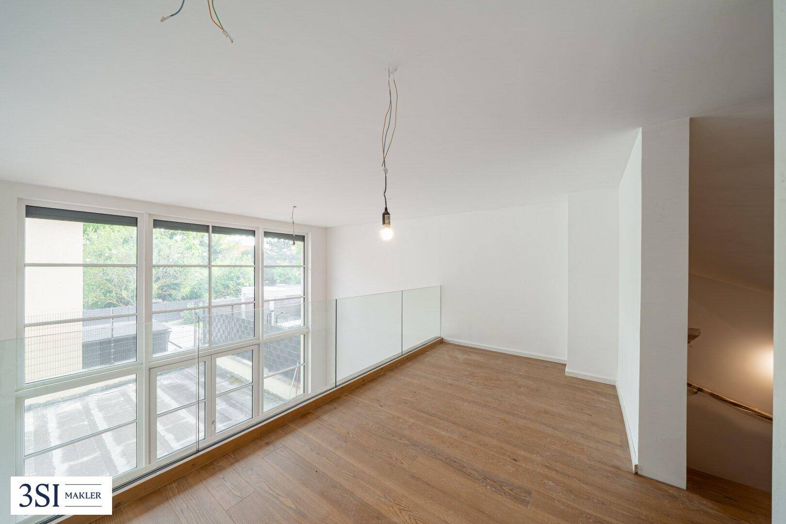 Heller Galeriebereich mit Glasgeländer, Holzboden und großen Fenstern mit Blick ins Grüne.
