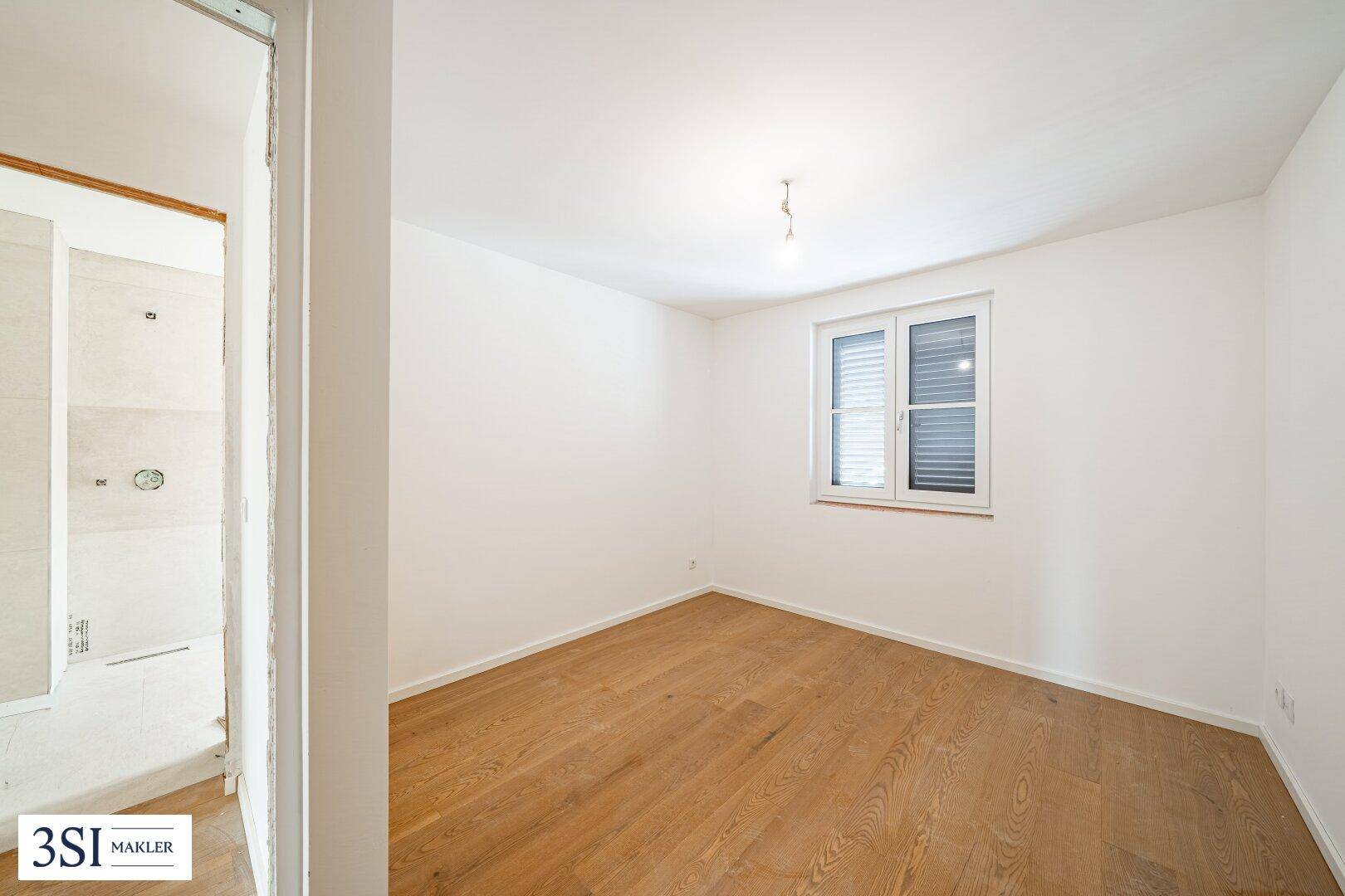 Unmöbliertes Schlafzimmer mit Holzboden, Fenster und Zugang zu einem Badezimmer.