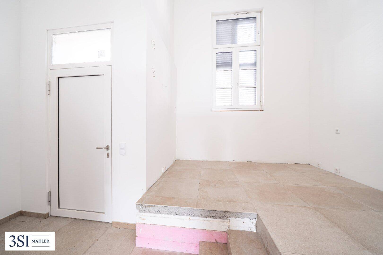 Unfertiger Innenraum mit gefliestem Boden und einem Fenster, bereit für den individuellen Ausbau.