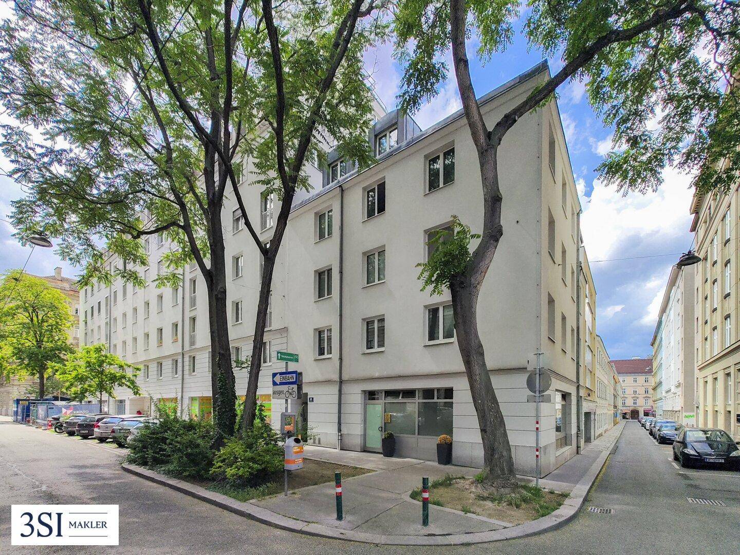Gebäude an einer Straßenecke mit Bäumen und parkenden Autos in einer städtischen Umgebung.