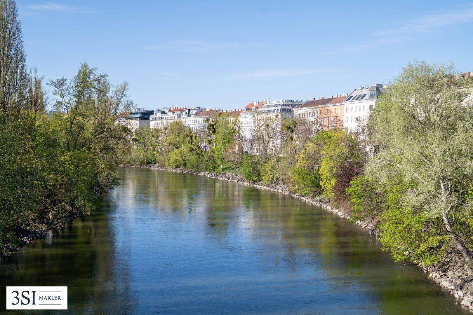 Flusslandschaft mit grünen Ufern und einer Reihe von Gebäuden im Hintergrund.