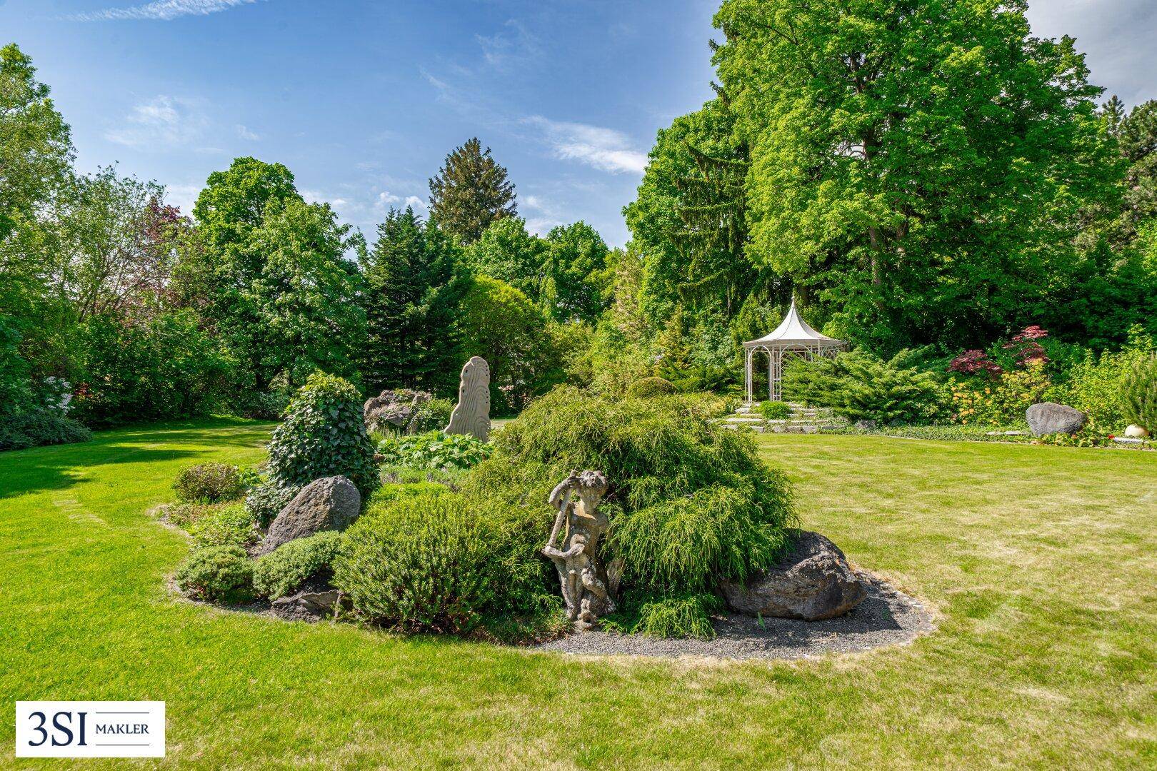 Weitläufiger, gepflegter Garten mit Ziersträuchern, Felsen und einem Pavillon im Hintergrund.