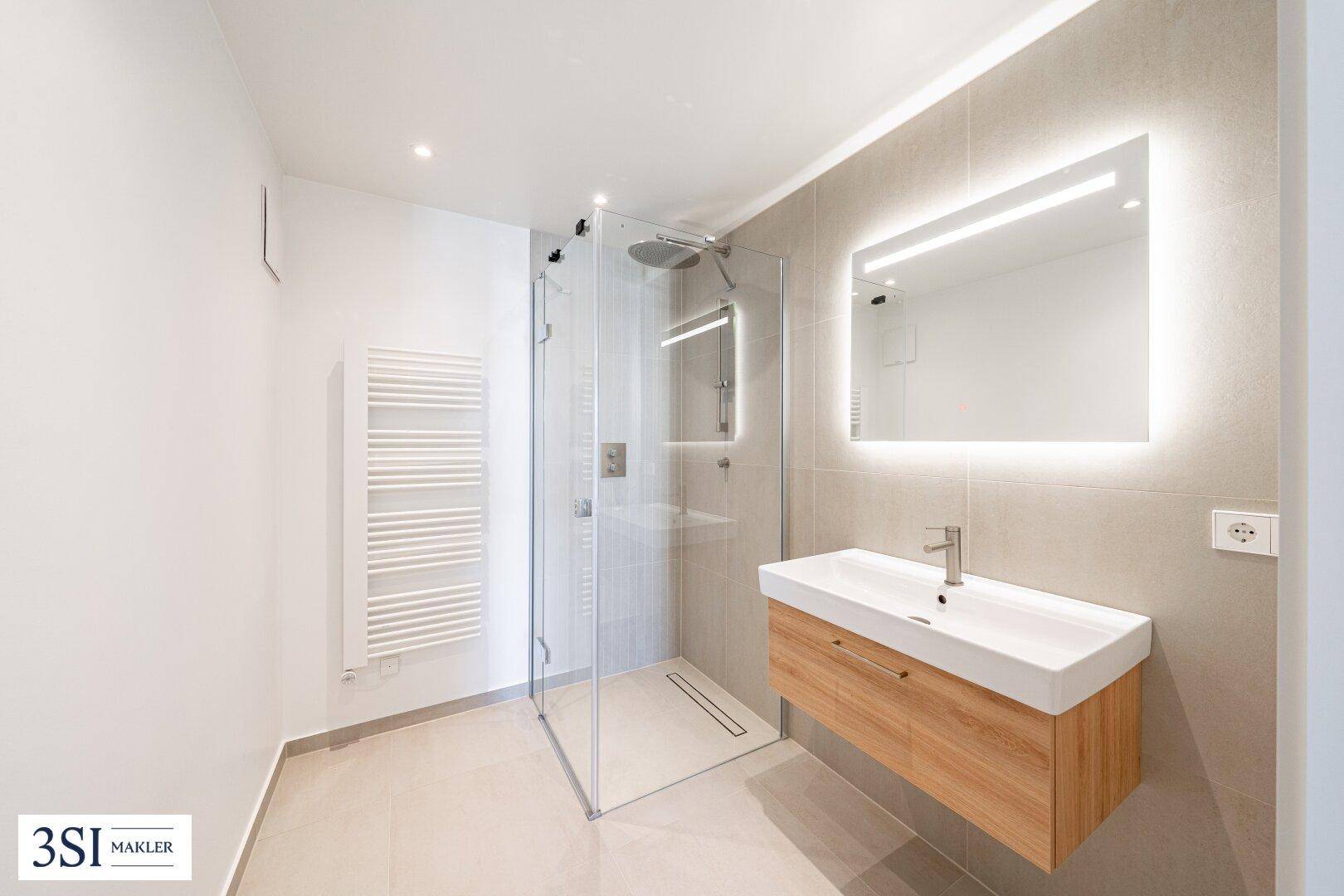 Modernes Badezimmer mit begehbarer Dusche, schwebendem Waschtisch und beleuchtetem Spiegel, in hellen Grautönen.