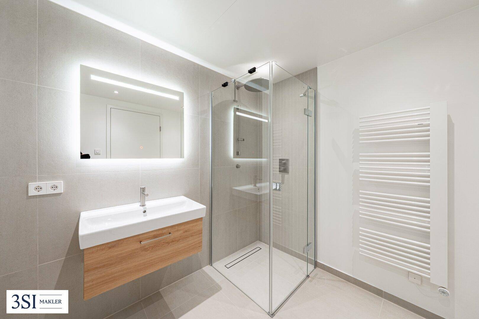 Modernes Badezimmer mit großer Glasdusche, Waschtisch und beleuchtetem Spiegel, in hellen Grautönen.