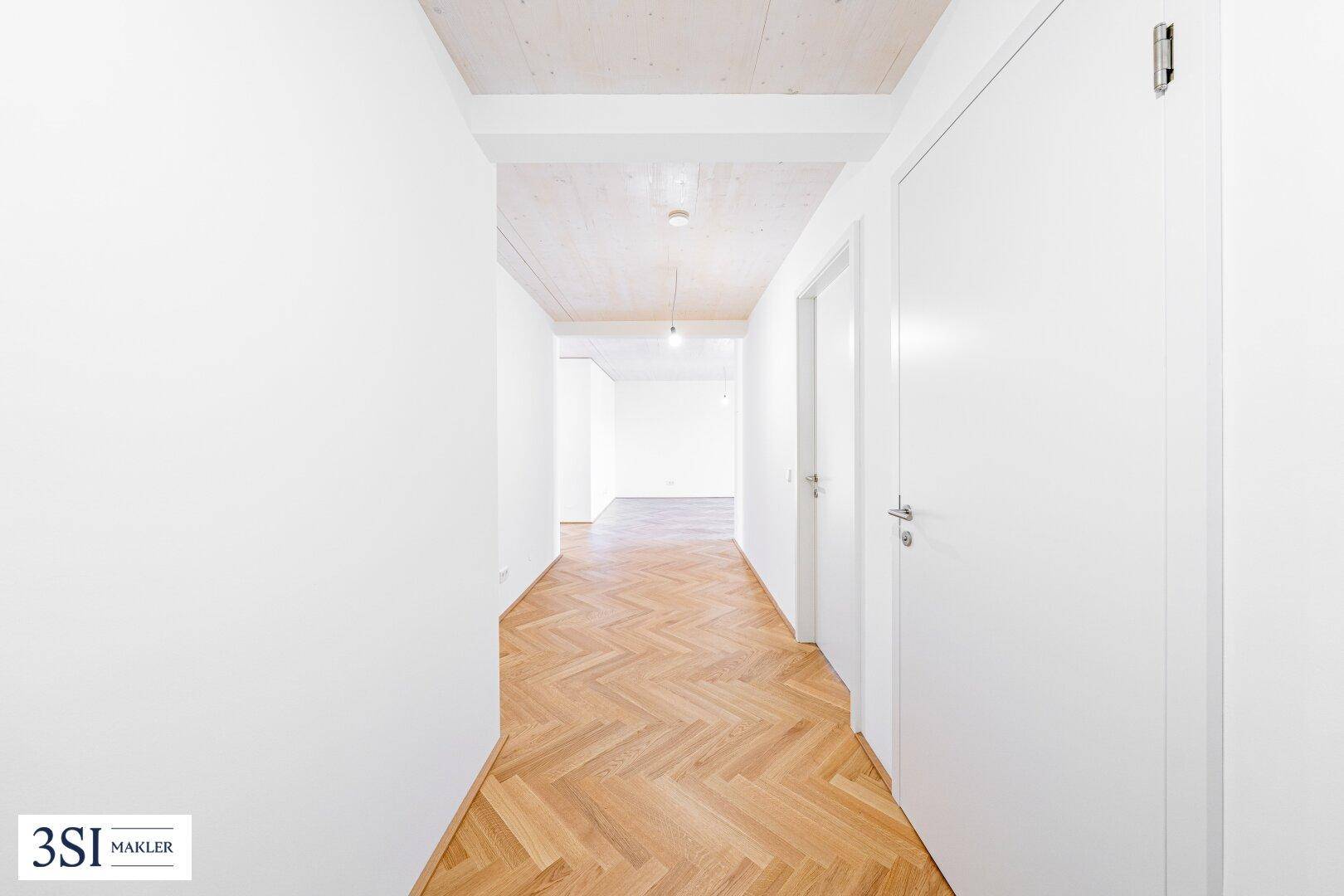 Heller Flur mit Fischgrätparkett, weißen Wänden und Holzbalkendecke, der zu weiteren Räumen führt.