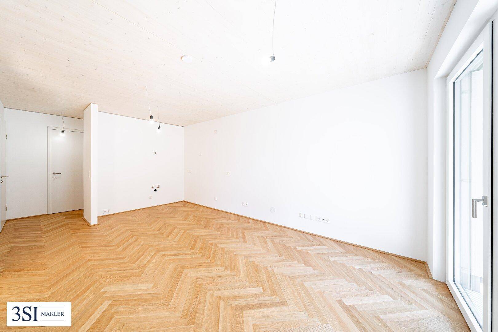 Geräumiges, unmöbliertes Zimmer mit hellem Fischgrätparkett und weiß gestrichenen Wänden.