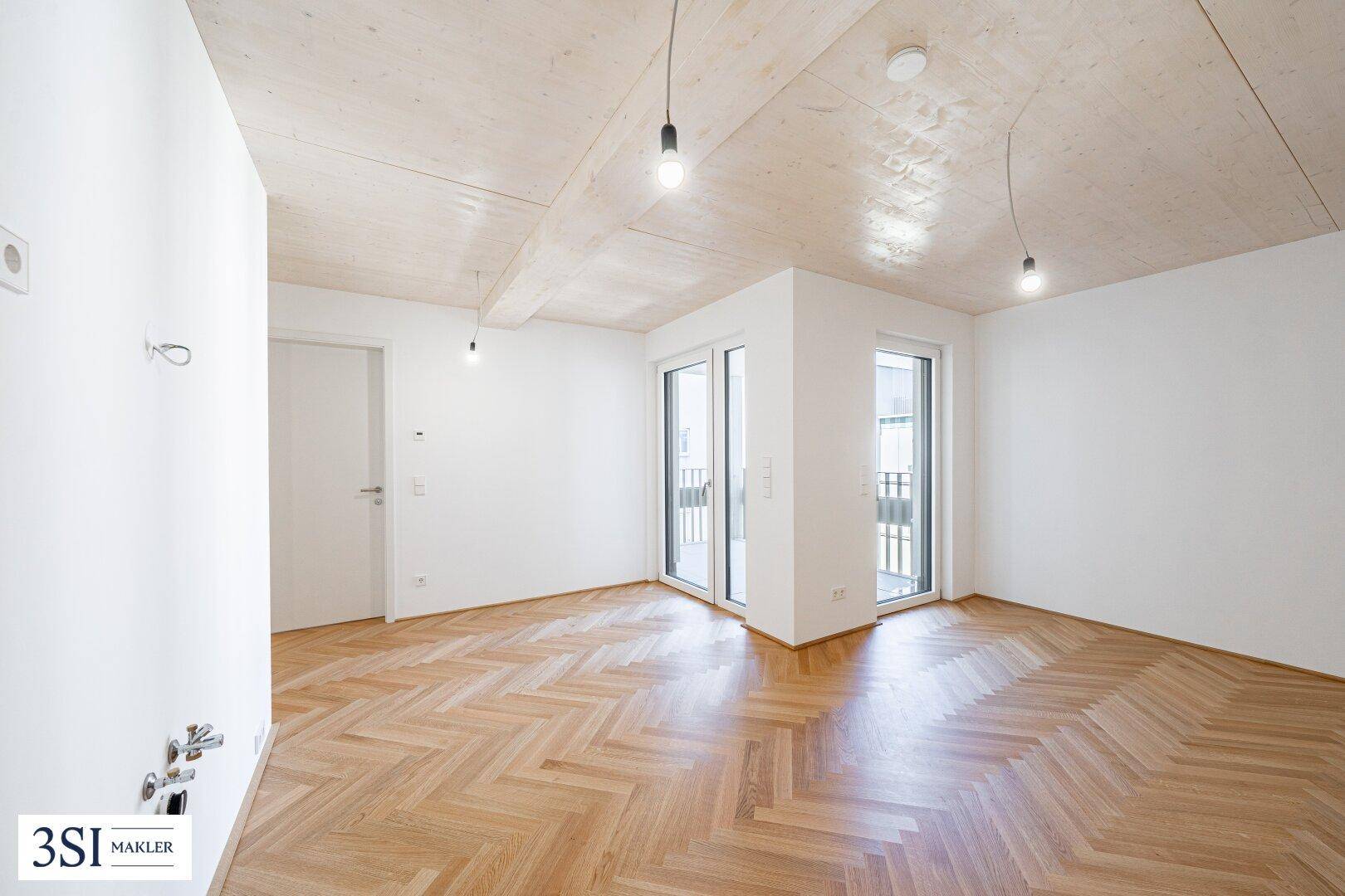 Heller Eckraum mit Fischgrätparkett, Holzbalkendecke und zwei Zugängen zu Balkonen.