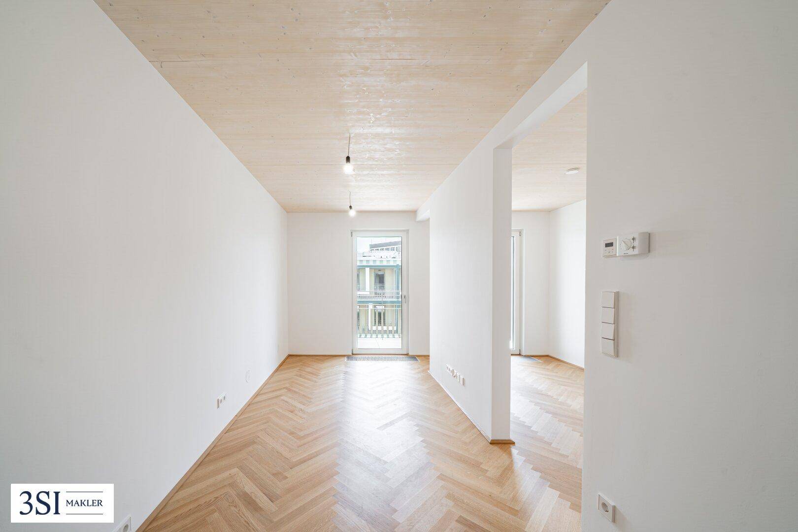 Heller Raum mit Fischgrätparkett, Holzbalkendecke und Zugang zu einem Balkon.