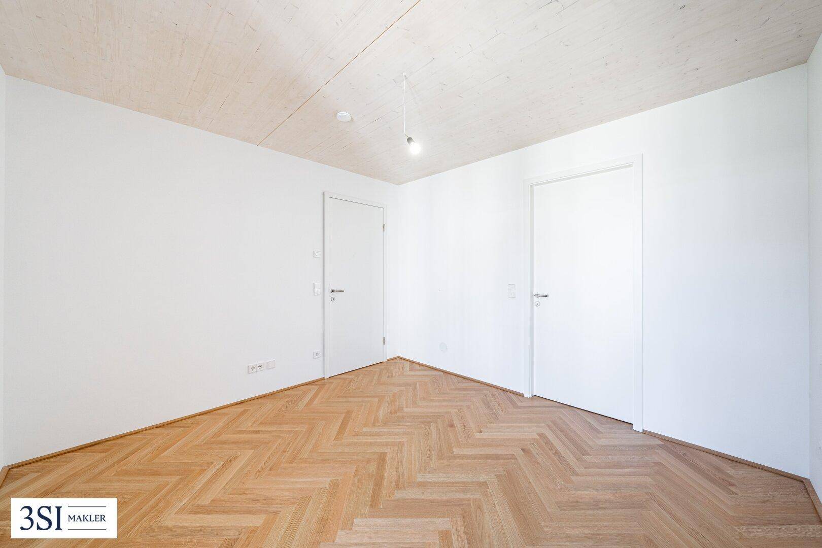 Helles, leeres Zimmer mit Fischgrätparkett und zwei Türen, ideal als Schlaf- oder Arbeitszimmer.