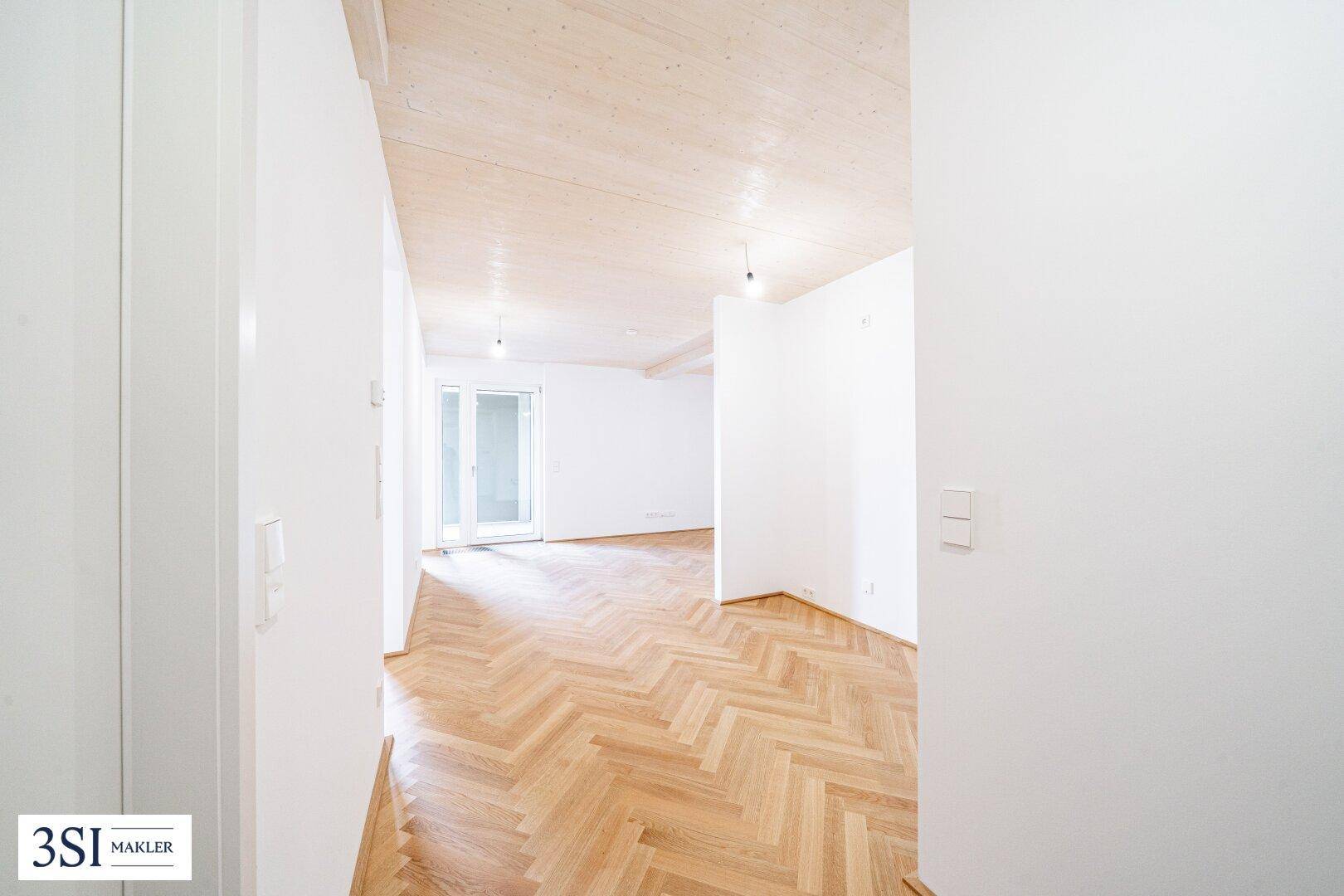 Geräumiger, heller Innenraum mit Fischgrätparkett und sichtbaren Holzbalken an der Decke.