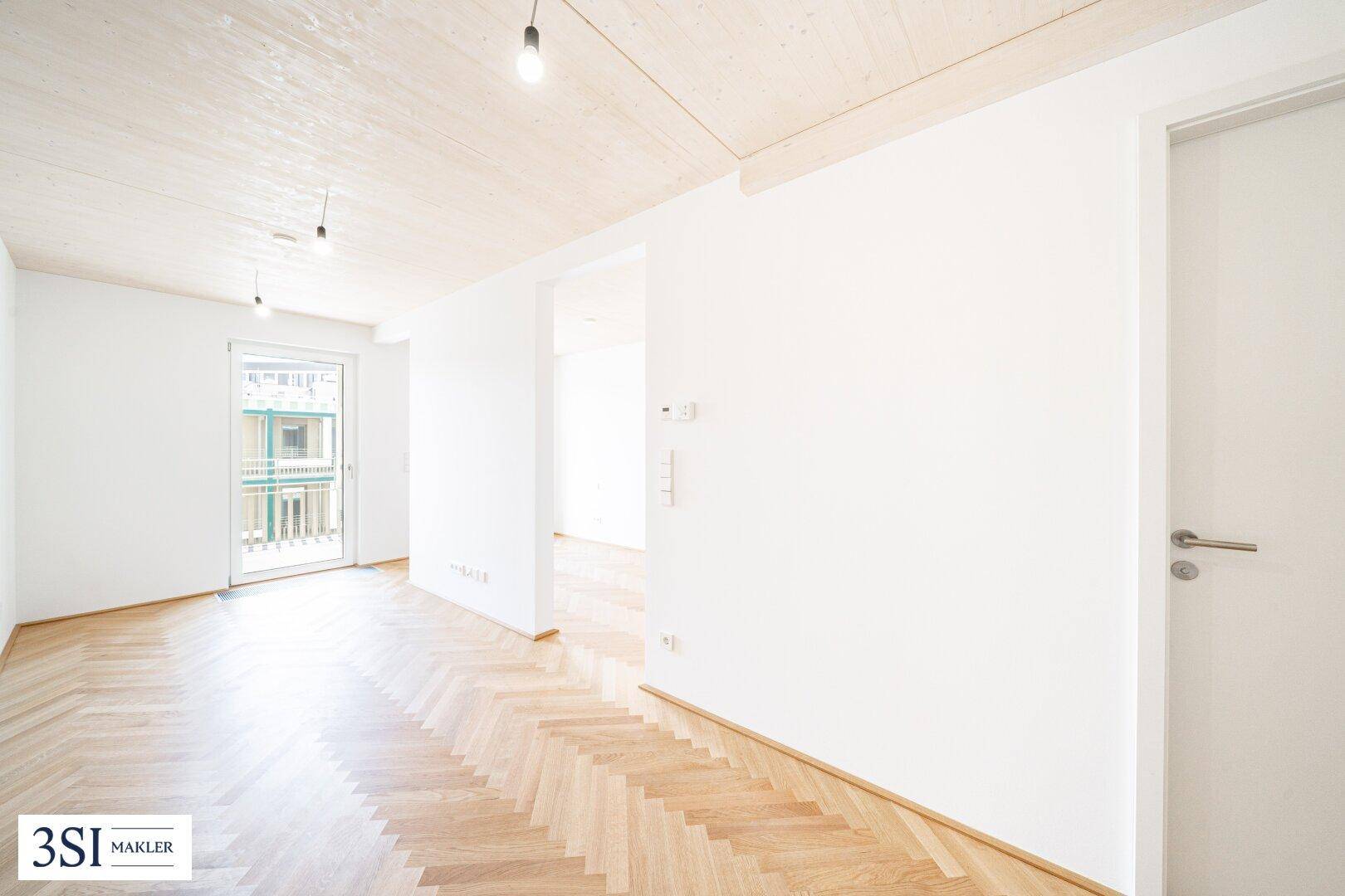 Lichtdurchfluteter Wohnraum mit Fischgrätparkett, weißen Wänden und Zugang zu einem französischen Balkon.