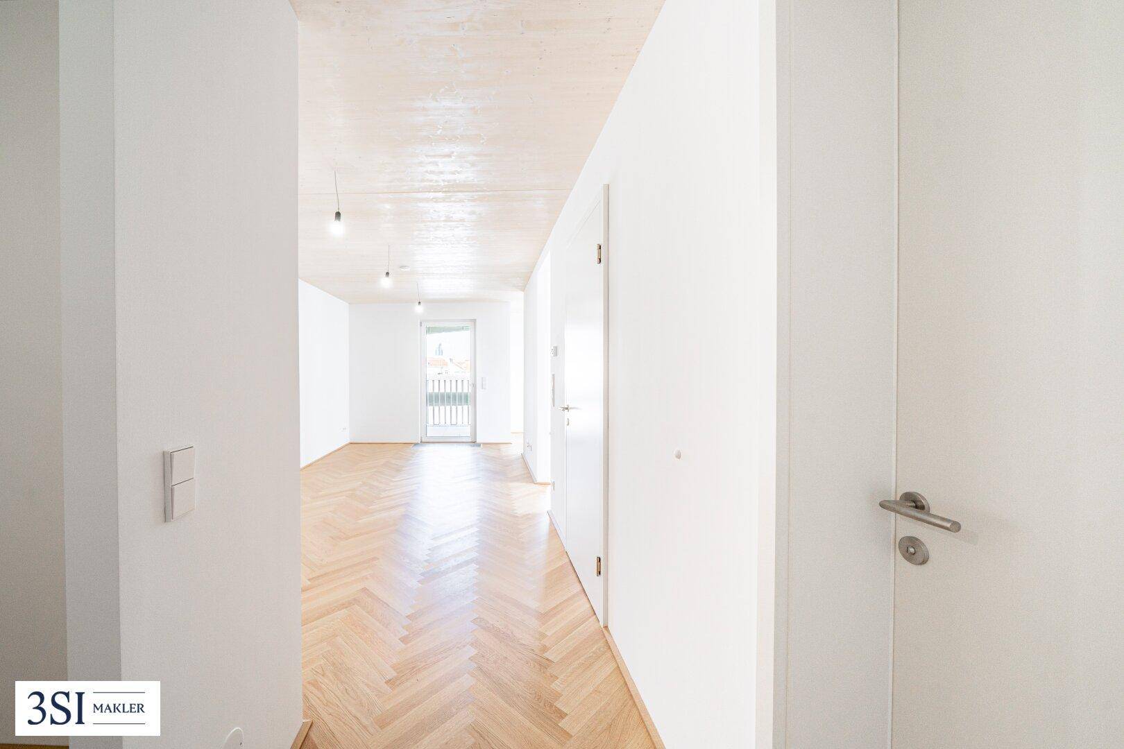 Langer, heller Flur mit Fischgrätparkett, der zu einem Zimmer mit französischem Balkon führt.