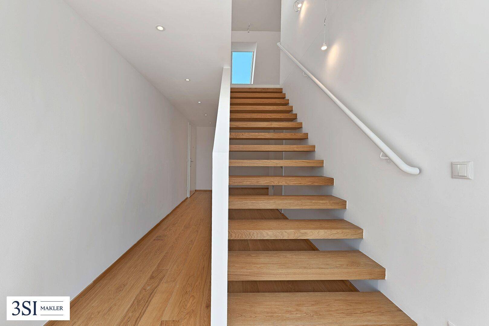 Heller Flur mit offener Holztreppe und Handlauf, der zu den oberen Etagen führt.