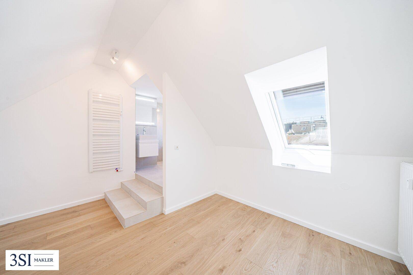 Helles Zimmer im Dachgeschoss mit Holzboden, Dachfenster und Blick in ein modernes Badezimmer.