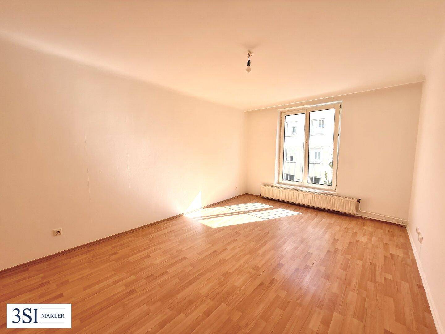 Leeres Schlafzimmer mit Holzboden und großem Fenster, das viel Sonnenlicht hereinlässt.