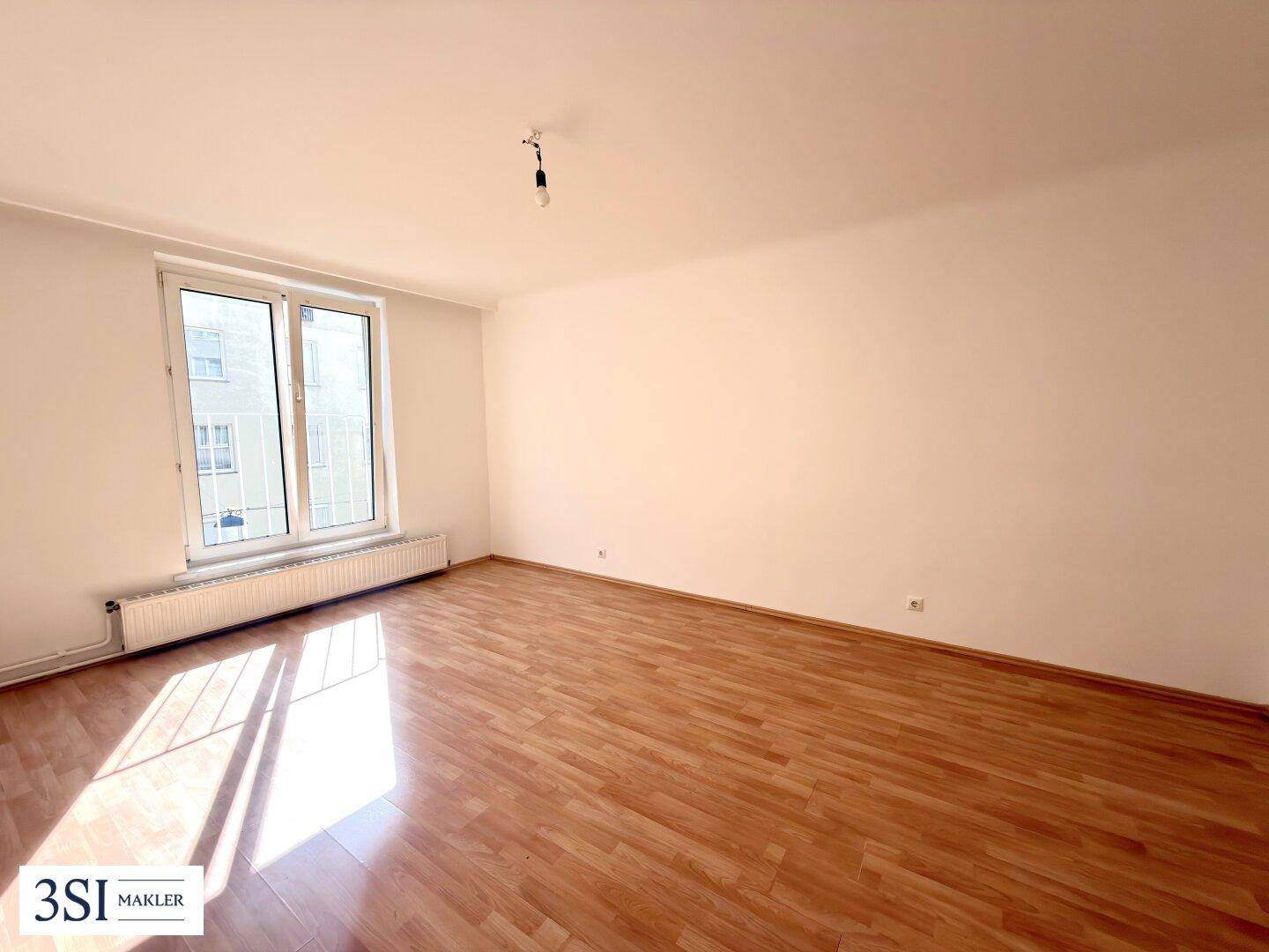 Leeres Wohnzimmer mit Holzboden und großem Fenster, das viel Sonnenlicht hereinlässt.